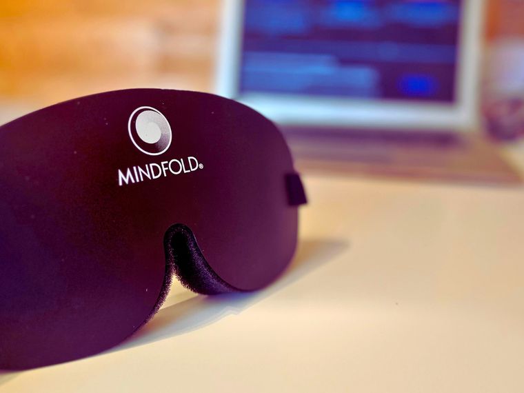 Antifaz opaco de la marca Mindfold apoyado sobre una mesa clara, con un portátil desenfocado al fondo mostrando una videollamada. Es el modelo de antifaz que se usó en todas las sesiones del estudio.